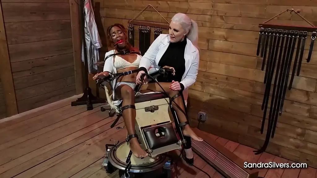 3516 Sandra Silvers and Victoria Ransom A Visit to Doctor Sandra Silvers' Sanitarium results in Bondage Orgasm Therapy for Beautiful Black Girl Victoria Ransom!<br><br>It was believed hysteria was caused by overstimulation of the mind. Unbeknownst to her, Victoria seems to be suffering from this condition! It’s clearly evident though - just take in how tightly strapped she is to a medical chair, how she hyperventilates behind her red ballgag, and her fidgeting form as she tries to figure out where she is! Luckily, Sandra is an expert in her field, called in to cure Victoria of these concerning symptoms…through a different form of overstimulation! Striding in, heels clacking and corseted waist swaying as she takes in her patient, Sandra dons a white medical coat and gets to work.<br><br>First things first… Victoria has no need for that tight white dress. For this particular treatment, every part of the patient must be exposed. Sandra smirks, cutting the dress away from her pleading patient, before adjusting the straps around Victoria’s legs. After all, closed legs a cure does not make. Spreading Victoria’s legs nice and wide, Sandra restraps her, ignoring the muffled whimpers. Trailing her nails over her patient’s body, Sandra inspects her. Whimpering in white lingerie now, Victoria squirms but it’s clear she’s securely strapped… Sandra’s silence only solidifies her helplessness!<br><br>Now… for treatment. Sandra slinks behind Victoria, rattling the metal medical tray and enjoying Victoria’s cries of consternation, before revealing a vibrator. This treatment is time tested…and may take a few trials to take hold! With another smirk, the vibrator is slid between the struggling patient’s thighs as Sandra expertly extricates wave after wave of whimpering orgasms. Victoria bucks and gasps, distraught as the pleasure overpowers her. This continues until Sandra is convinced her patient is close to cured. She returns the vibrator to the tray, leans Victoria’s medical chair back with an ominous clank, and leaves the room… leaving her patient to wonder if she’ll ever be free…