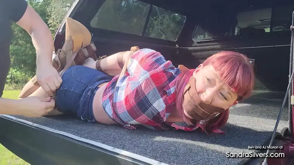 3532 AJ Marion and Ben " Well, ain't this a wild rodeo, y'all? Here I am, little ol' me, Alice Jane Marion, in my red, white, and blue plaid shirt huggin' my curves just right, them denim shorts ridin' high on my pantyhose-d thighs, and my trusty brown cowboy boots planted firm as a buckin' bronco. That broad shouldered varmint's got me from behind, his strong arms wrappin' me up like a lasso, tilin' my head back so I can't help but gasp through this leather panel ballgag he's slapped on my mouth - oh, it's mufflin' my sassy yelps somethin' fierce, makin' me all squirmy and helpless against his grip. My red hair's a mess of wild waves, and with my hands tied up front, I feel that thrill of bein' caught like a feisty filly in heat, my body buzzin' with that naughty edge that makes a gal like me wanna kick up some dust and play rough.<br><br>Now, that roughneck's draggin' me away, tossin' me into the bed of his truck like I'm his prize calf at the county fair, still gagged and wrigglin' against these darn restraints that got me feelin' all kinds of exposed and achin'. The truck bounces over them rugged country roads, my cowboy boots scrapin' against the bed-rug floor, sendin' sparks of static electric friction up my legs as the open windows whip wind through my red hair, leavin' me at his mercy out in the open. Every jolt hits my body just right, makin' me whimper and squirm, my thoughts racin' with what he might do next - will he tease me more, or let me break free for a chase? It's a bumpy ride that'll leave a gal's heart poundin' and her boots dirty, but darn if it ain't excitin'. "