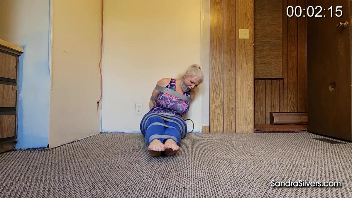 2988 Sandra Silvers &amp; Amanda Foxx Tightly Taped Tied Two in the Trailer Team Up to Attempt Escape!<br><br>With only tight leggings covering her round ass and a spandex sports vest over her huge boobs, Sandra has been cruelly duct tape tied and gagged and left in the back bedroom of a broken down trailer home. With the silver tape holding in mouth-stuffing and wrapping the circumference of her head, cruelly cleave gagging between her lips, she can’t call for help. She can’t do more than muffled mmphs as her gym going girlfriend Amanda is brought into the tiny room and stashed next to Sandra on the dusty, dirty carpet.  Their only chance to escape is to free therselves from the tape torment, but that is unlikely to happen with wrists tightly taped together behind their backs. Once they hear the trailer door close Sandra struggles about the floor to reach scissors left on a high up table top. Amanda tries in vain to reach the implement of their escape with her long legs, but neither can reach… Till Sandra manages to hold her curvy body high enough to grip those shears between her cleave gagged lips! What a feat… the barefoot pair unwrap themselves and each other from the super sticky sealing surrounding the daring damsels, running from the room, but it looks like the yoga toned two will be going nowhere fast after all...