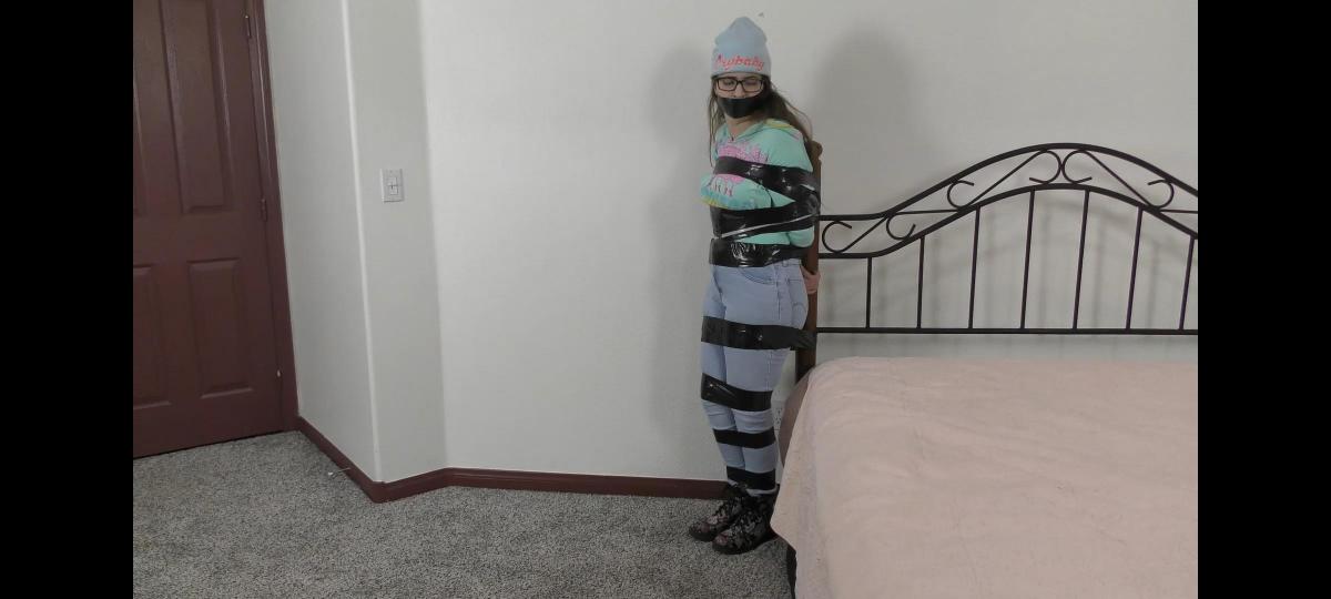 QT0983 Mia Nelson (is-qt-mn071) Poor Mia Nelson has been captured & thoroughly bound & gagged with duct tape. The video opens with Mia escaping out of the bedroom & scooching her bound body through the living room & down the hall toward the front door. It takes quite an effort, but Mia finally makes it to the door and tries to open it with her bound feet. As she fumbles with the handle, her captor sneaks up on her, hand gags her and pulls her back into the bedroom where she is taped standing to the bed post. Mia struggles, but cannot break the tape. Undeterred, Mia decides to yank the entire bed with her! This is a great struggle video with Mia looking adorable in her blue jeans, hoodie top, knit hat and eye glasses.