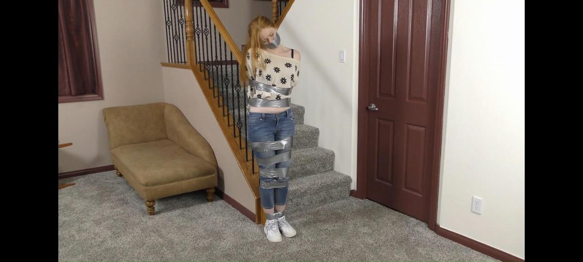QT0979 Celeste Evergreen (is-qt-ce011) Celeste Evergreen returns home to find her place in disarray – she has been robbed! Worse, the burglar is still there & despite her protests, Celeste ends up tape tied tied and gagged. But the feisty redhead isn’t about to give up as she vigorously tries to free herself before deciding to try and escape by scooching her way across the floor. She makes it to the door and almost has it open when she is dragged back into the room and more thoroughly tape-tied standing to the stair banister. A great high energy performance by Celeste!