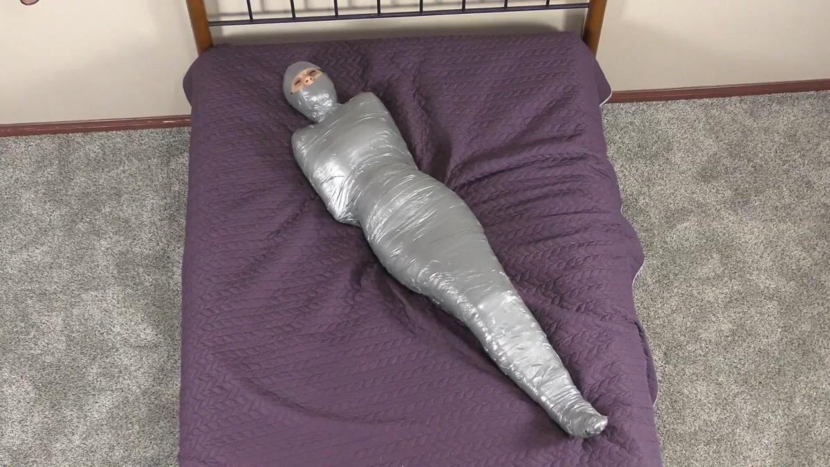 QT1103 Kristen Moore (is-qt-km062) n one of the strictest mummifications we have ever done, Kristen Moore is captured, has her wrists & ankles bound with rope and is then wrapped from head to toe in vet wrap, including having her neck, head and feet wrapped! She struggles fruitlessly on the bed before a complete layer of silver duct tape is added over the vet wrap, also a full head to toe wrap. A two layer mummification from which Kristen has no hope of escaping!