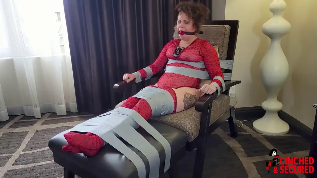 1407 - Bailey Taped, Gagged, Drooling and Helpless Bailey and I were supposed to have a relaxing weekend getaway, but it seems she decided that hanging with her friends at the resort was more important than spending time with me.  She's going to learn ALL about staying in now, whether she likes it or not.  I taped her tightly to a chair, stretched her legs out straight, and taped those to the footstool. She could life her legs with effort, but there was no way she could hold them for long with the added weight of the furniture she was welded to!  Then I taught her about my idea of quiet with a big fat ball gag!  Now I could watch her moan, drool and gurgle her dismay at her plight! Of course, I always like to make things worse, so not only did she have to put up with the feeling of her saliva running down her chin and neck and slowly dripping into her top and down her chest and breasts, she got to do it in total darkness thanks to the blindfold I added! A little electrical tape anchored her hands flat and tight against the arm of the chair, ensuring she would not more at all!  Ya know, this arrangement is so nice, I just might extend our stay a few days!