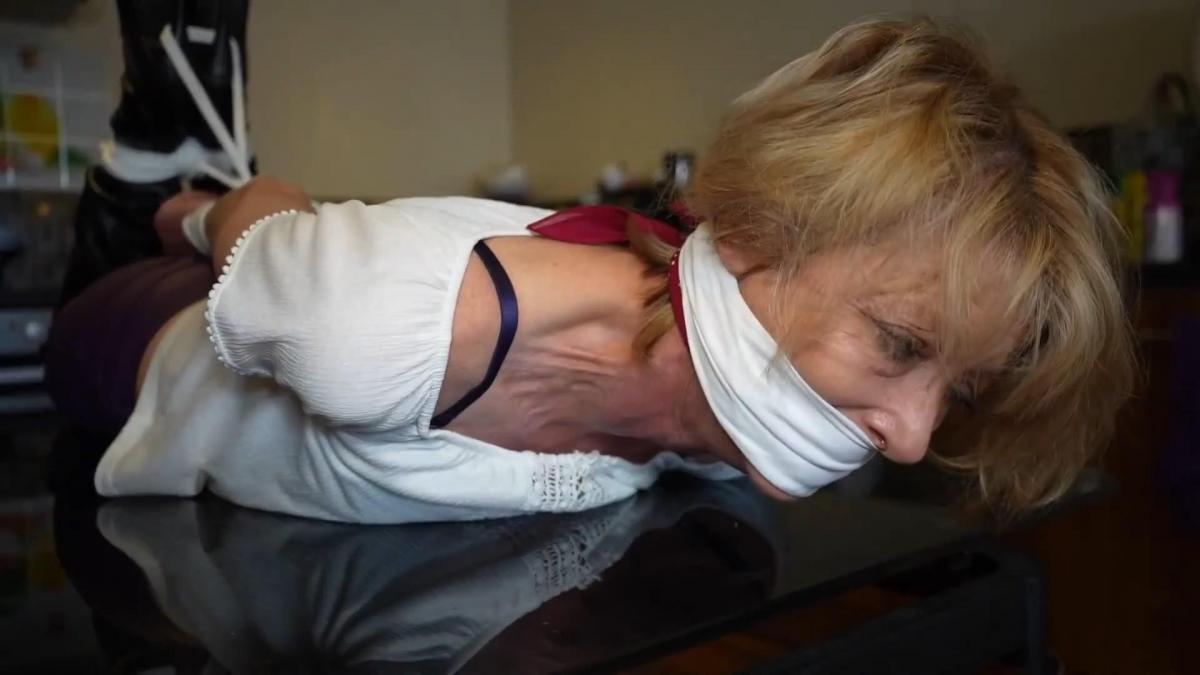 The milf nabber, featuring toni g Toni g finds herself at the hands of the milf nabber. She's been dragged to hes apartment with her wrists bound behind her back and a nice tight wide bandana cleave gag between her lips. The nabber sits her her and begins tying the milfs ankles together before leaving her to struggle. She rolls round the floor in an attempt to free herself but the ropes are too tight, her crys muffled by the scarf between her lips. She drags herself to the wall and takes in her surroundings before heading g to the sofa. She crawls across the floor and hoists herself up. She once again trys to reach the rope at her ankles but they are just out of reach. Unsure if the man is still around She gets to her feet and grabs a chair from close by for support and hops across to the door in which she was brought. She manages to get to the door bit unfortunately for her the man was listening in to all her struggles and was waiting for her from behind the door. The man grabs the gilf and drags her to the catches table where he proceeds to hog tie her. Now totally helpless and sitting high poor toni has no chance of getting free. The ropes now pulling tight at her wrists and ankles. All toni can do now is squirm and sob through her gag. To make things harder for toni the man then pulls out a handkerchief and gives her an over the mouth classic gag to silence her screams for help more. He leaves and now poor toni is tightly bound, hogtied and gagged in her leather pants and knee high boots on the kitchen table where she awates her fate.