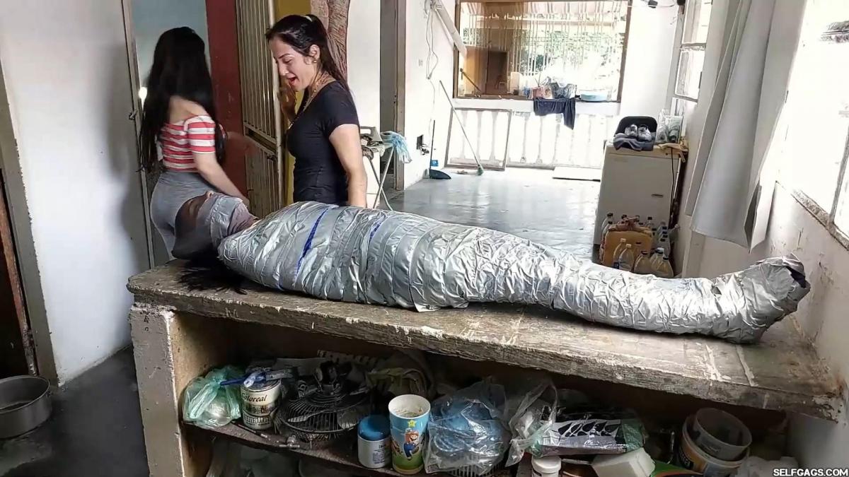Wrapped Up And Stored In The Attic Maria Martinez is bound, tape gagged, and struggling on the floor. Two women enter and put her on a chair, replacing her classic tape gag with a handgag before tightly gagging her with a dirty sock and duct tape. The women then put her in a sleepsack, tape mummify her, and carry her to the attic where they encase her head in nylons and lay her on a counter. Maria is helpless and struggling throughout the ordeal!