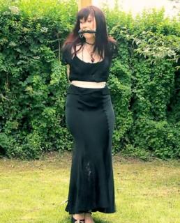 Garden Ornament Syrah Lee is tied to a pergola post outdoors. Her hands are bound behind her back, and she's secured further at the waist and at the ankles. A large bitgag is lodged in her mouth. She is dressed in a long, elegant black dress.