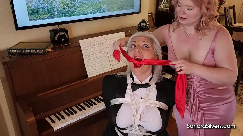 3394 Sandra Silvers &amp; Ami Mercury Piano maestro Sandra is dressed to the nines for a recital with an audience of one, her live in MILF lover, Miss Mercury... In a sharp tuxedo, Ms. Silvers sits at the piano in her parlour and begins to play, but she doesn't get very far through her repertoire before Ms Mercury, in a long, low-cut ball-gown, decides to spice things up with some rope bondage. Sandra soon finds herself trussed up along the length of her body: legs bound in several places, another rope between them, more bonds around her magnificent chest, wrists lashed together behind her - totally helpless! She squirms delightedly, tossing her hair, smiling broadly, as Ami fondles her breasts. Then it's time for a gag. Sandra's lips are parted by a red silk scarf cleave, muting any protests as she's groped some more. While our bondagette's breasts strain against her tight formal shirt; by way of contrast,we see a lot of her captor's fine cleavage as she teases and taunts...<br><br>Taking pity on her struggling captive, Ami takes up a watching brief from across the room, observing Sandra moving from piano stool to the floor, sitting, standing, then lying down, half-heartedly trying to get free. After a while our sexy domme finds some more rope, and proceeds to hog-tie the pretty performer, ahead of more hands-on hi-jinks, to the obvious pleasure of her target. Finally, with both phases of the performance over, Ms Mercury takes her leave. The bound beauty will have to get free on her own...