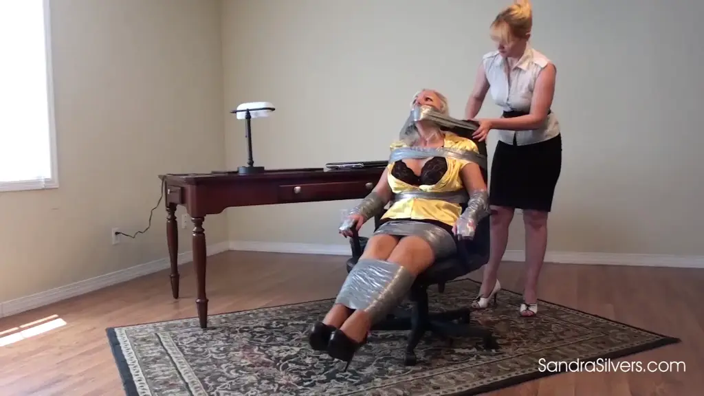 1591 - Sandra Silvers &amp; Lisa Harlotte Lisa used an entire roll of duct tape was used to pin Sandra to the office chair. Her long, curvy legs are taped together and her forearms and fingers are tightly tethered to the arms of the chair by the sticky restraints!  Lisa isn’t done with Sandra yet.  An entire bandanna is stuffed into Sandra’s mouth and behind her teeth to keep her silent as several strips of duct tape strategically keep it in.  Deciding that the strips are not enough, Lisa wraps the duct tape all the way around Sandra’s head, over and over, making absolutely sure her captive stays silent. More tape is used to hold Sandra’s shoulders to the chair and then even MORE tape is employed to immobilize Sandra’s head.  Lisa leaves her prey to struggle and Sandra wonders how can she now possibly get someone to help her? Then she spies the cell phone on the desk…