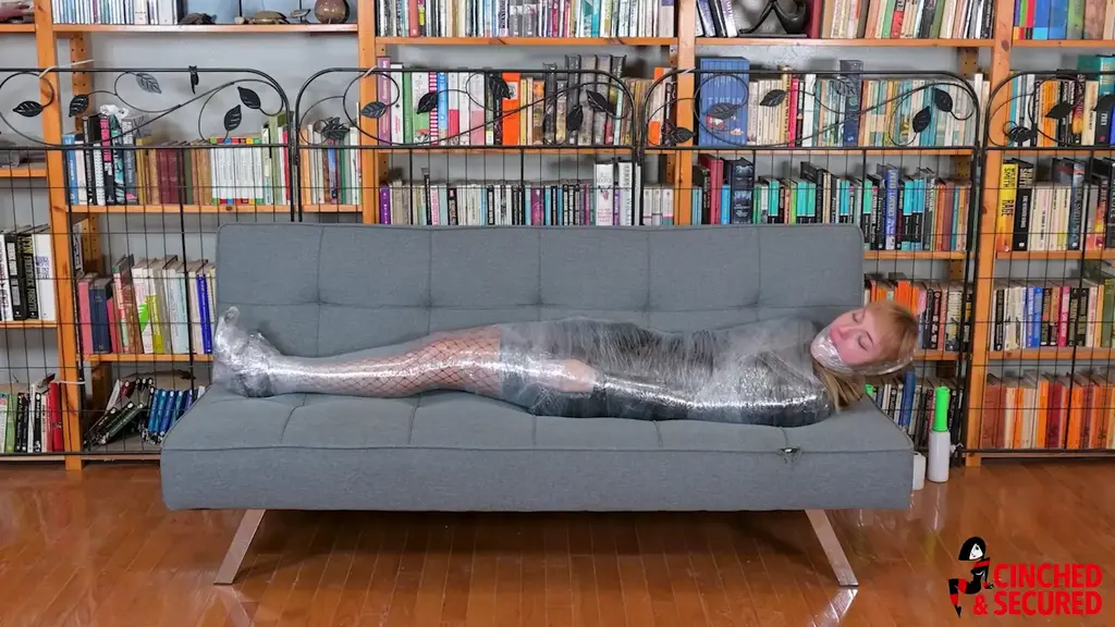 Cara Day - Cocooned and Gagged in Latex and Plastic Wrap Cara loved the feel of her new latex mini dress! In fact, she wanted more than simply feeling it on her skin. She wanted to be constricted in it, crushed, encased. She sat wrapping plastic wrap around her hand, trapping her fingers into tight little balls. The feeling only fueled her desires, and she asked her friend to help her achieve maximum compression, and he was only too happy to oblige. He wrapped her entire body, from neck to feet, fusing her arms to her sides, turning her legs into one solid column incapable of even bending, and even wrapping her heels to her feet, preventing her from taking even the smallest steps. Finally, he popped a ball gag into her eagerly waiting mouth and propped her standing in the corner. She writhed and moaned, her eyes rolling back in ecstasy as she squirmed and teetered on her fused feet. Her friend, however, decided she needed even more and wrapped the plastic around her face. She stared in shock, unable to speak, and he reversed the path of the plastic, winding it under her chin and over her head again and again. She was now silenced, and he dragged the poor girl to the center of the room, Cara taking pitiful tiny hops to remain upright.  She would not stay that way for long, however, as her friend trapped her on her side on the sofa! Completely helpless, Cara rued the moment she had enlisted her friend, and wondered just how long she would remain trapped in her plastic prison!