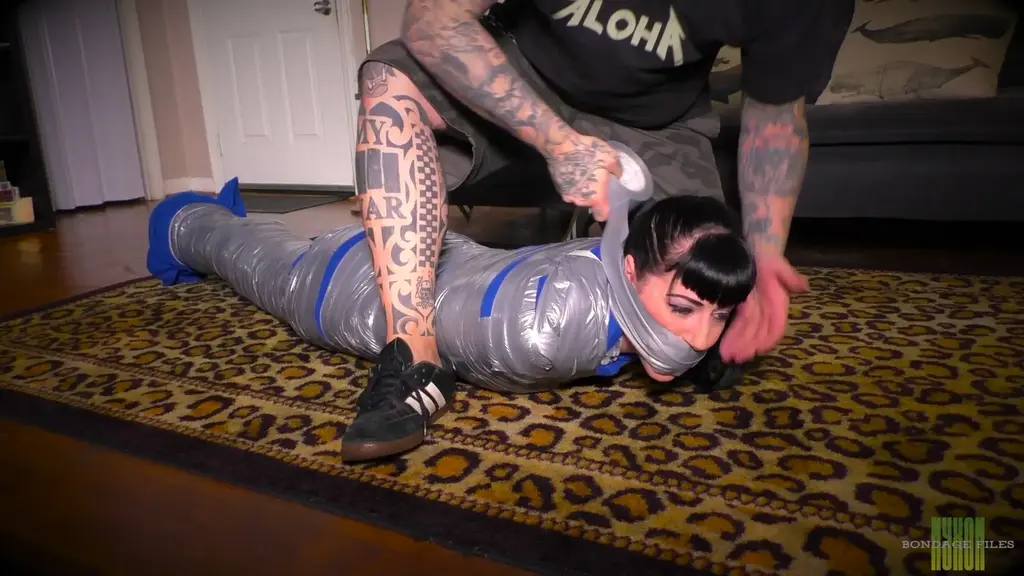 Nyxon Cat Burglar Caught &amp; Punished With Mummification Cat burglar Nyxon has just broken into a house, and is beginning to snoop around. Suddenly, the owner of the house appears & demands to know what Nyxon is doing in his house. Nyxon stammers searching for something to say, but the man grabs her & starts wrapping duct tape around her securing her arms & her legs. He wraps her up in a fuzzy grey blanket, pushes her up against the door, & starts to tightly wrap her in duct tape. Next, he takes a blue fleece blanket, wraps it around the grey one & tightly wraps her in duct tape from her shoulders to her ankles. He then lowers her onto the floor, takes a large pair of white panties, shoves them into her mouth, and then wraps duct tape around her head. He then takes a large tube sock, ties that around her head & wraps even more duct tape over that. He then leaves her there to struggle on the floor in her very tight mummification.