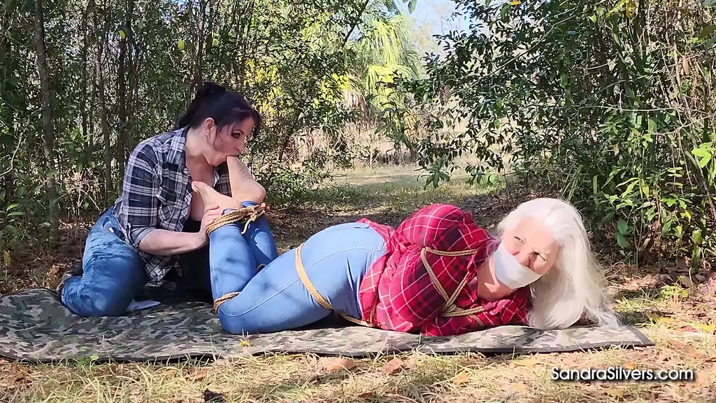 3650 Sandra Silvers, Caroline Pierce, and Ben Hot Footed to Fettered Foot Worship: Silver-Haired MILF Sandra Snatched by Caroline, Hopped Outdoors, Hogtied by Toe Licking Lesbian, and Boot Burgled by Ben!<br><br>Sun-scorched scrub of the Florida countryside sees silver-haired MILF Sandra Silvers strictly secured and shamefully shoved, hopping across the sandy soil. Clad in tight blue denim jeans that grip her thick thighs and hourglass hips, under a red plaid flannel shirt straining to contain her substantial, pendulous breasts, Sandra is hopelessly hobbled by heavy coils of natural brown jute rope. The rough fiber bites into her bountiful bosom, framing her large, tanned breasts and slightly erect nipples in a chest harness that leaves her cleavage crammed against the lumberjack print fabric. Her arms are wrenched behind her back and welded together with unforgiving knots, while her ankles are lashed tight, forcing her to hop helplessly through the dry grass.<br><br>Dominant diva Caroline Pierce, her dark locks lashed in a severe high ponytail, guides her gagged captive to a secluded, shaded spot beneath moss-draped trees. Sandra’s whimpers are perpetually pacified by a pristine white vet wrap gag pulled taut across her lips, sealing her speech as securely as the rope seals her limbs. Caroline commands the curvy captive to settle upon a camouflage-print blanket spread across the leaf-littered ground. Sandra sits, submissive and silent, her tan hiking boots planted in the dirt, anticipating the inevitable exposure.<br><br>With sadistic slowness, Caroline captures Sandra’s thick white socks, peeling them from her sweaty soles after unlacing and removing the sturdy boots. The dominant mistress presses the damp, stinky socks to her nose, inhaling the pungent, musky scent of Sandra’s stroll through the scrub before turning her attention to bare, vulnerable feet. Caroline cradles the captives’ high-arched soles, worshipping the wrinkled textures and pale pink heel pads with her lips and tongue, savoring the salty sweat and natural funk of the fettered feet.