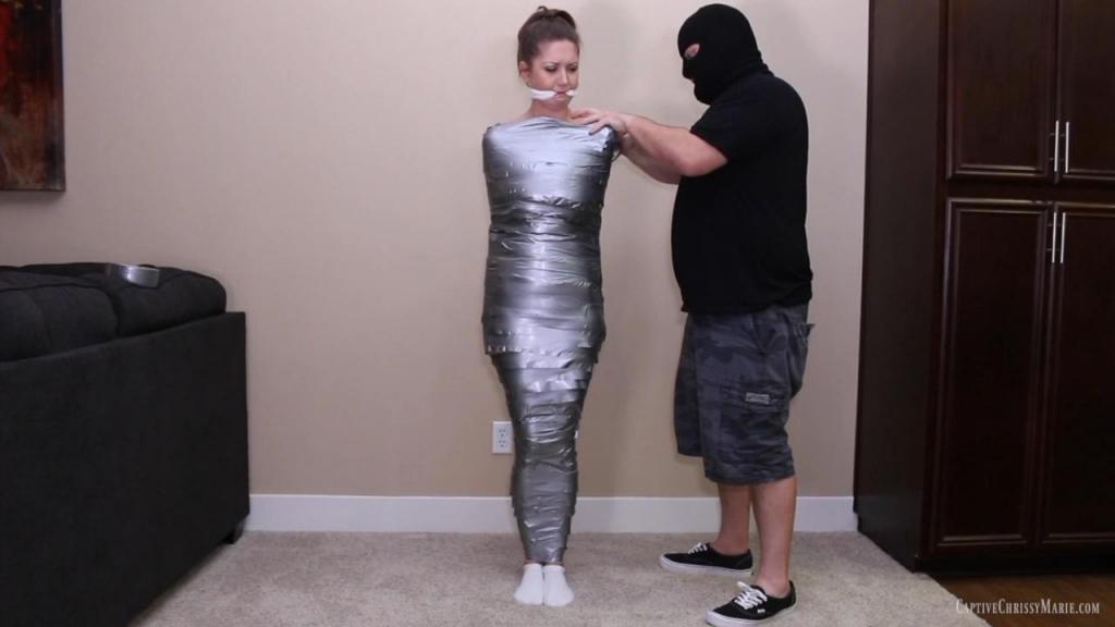 0191 Wrapped &amp; Gagged For Shipment Chrissy was out on a run when she suddenly realized someone was following her! She tried to run faster to get home but she was already too tired and he caught up to her!  and her taped her just enough to secure her for now while she takes a nice ride in his trunk! He brought her back to his hideout where he left her off while he notified his boss of the new merchandise. <br><br>Part one begins as she wakes up and groggily moans at her throbbing headache. She starts to notice that she can barely move her arms since they are pinned down with duct tape! Then she starts to remember being followed while she was running, and realizes she must be in imminent danger. She struggles against the bonds but she is far too late, he has already heard her wake up. The man enters the room armed with a white cloth to silence her distressed pleas. He pulls it tightly between her teeth, cleave gagging her as she goes wide eyed in shock and confusion. She has no idea what he wants from her! Next he shows two rolls of tape and she doesn't understand, he already taped her up! She begs for him please don't me up more! If only she knew what he has in store for her!<br><br>Starting from her ankles her wraps the sticky tape around every bare inch of her naked body and she really starts to panic now. She begs him to stop, he is really starting to scare her! He sure as well can't do that, today is shipping day and he needs to make sure she is secured in the most efficient and compact way for transit to her new owner! She can't believe her ears when he breaks the news to her. Half frustrated and half fearful, she wiggles and whimpers while trying to maintain her balance until she is fully mummified from her neck to her ankles! Her puts her the floor and we see that she really can barely wiggle or move at all! He tells her shipping is in 30 minutes and leaves her to struggle, but she cries out for him not to leave her like this! She ends up being too loud so he comes back and decides he needs to tape her mouth shut over her cleave gag. Once she is quite efficiently double gagged he leaves her to struggle yet again while he makes sure plans are in order. She has never felt so constrained! She is so desperate to find a way out but the only thing she manages to make it out of is her socks! Now barefoot her soles wrinkle as she fights against the tightly wrapped tape, soon realizing that she won't possibly escape! She mmmppphhs and struggles in peril waiting for her inevitable fate!<br><br>