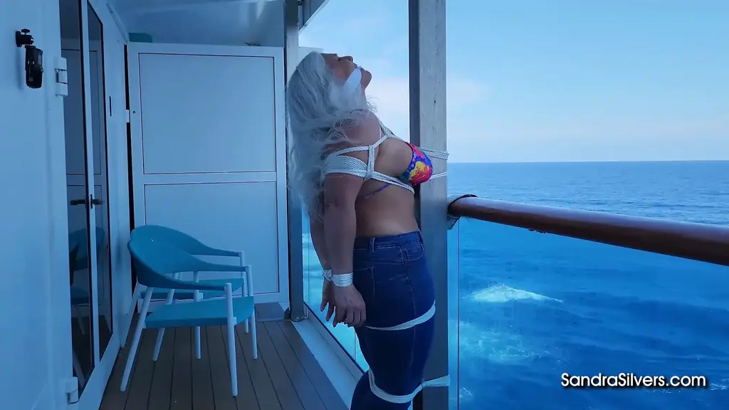 3730: Sandra Silvers and Ben with Catherine Sterling Cameo On the open-air balcony of a luxury cruise liner, curvy confined Sandra Silvers stands strapped in a straining rainbow bikini top and skin-tight blue denim jeans, her voluptuous figure lashed lewdly to a cold metal pole with coils of cruel white rope cinching her swelling breasts, circling her thighs, and pinning her bare ankles together as her toes, painted, point in peril upon the wooden deck. Ben has delivered this barefoot, busty broad to the waiting Catherine Sterling - who watches with wicked delight as rope after rope is added to captivate this MILF further in a pole tied predicament - from her POV perch as the ocean wind whips Sandra's silver hair across her cleave-gagged, grimacing face, her glistening bare soles flexing and her fettered feet fighting against biting binds while her bound breasts bounce and heave with every helpless struggle against the vertical pole overlooking the endless blue waves. Catherine's own flirtatious bare toes wriggle in the foreground of the frame as she savors the spectacle of Sandra's seaside suffering, the cameo cutie having orchestrated this entire onboard emergency evacuation solely to secure this sublime, submissive, seaside show of bondage torment.
