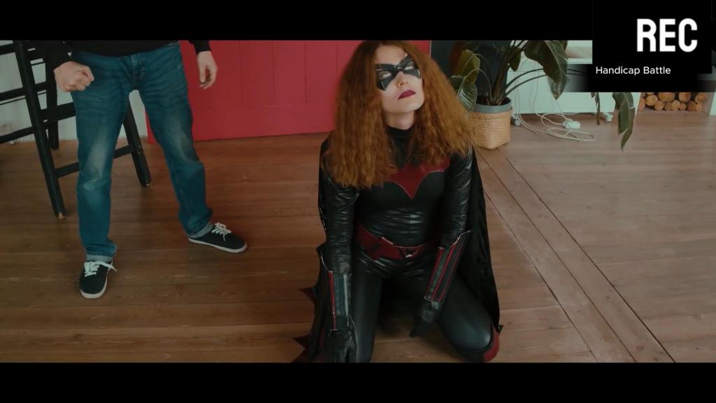 Handicap Battle A superheroine is defeated by her enemy in a latex peril scenario involving hair-pulling, unmasking, de-booting, bear hugs, and rough male handling.